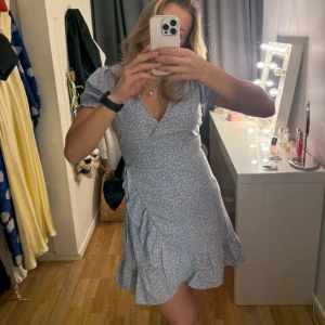 Blå omlottklänning med blommigt mönster - Blå blommig klänning från Nelly, använd en gång och som ny!