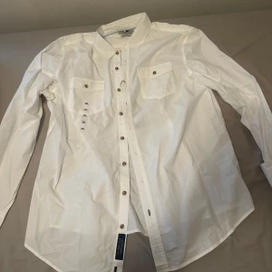 Vit skjorta med knappar - Säljer en stilren vit skjorta med knappar och två bröstfickor. Skjortan har en klassisk design med långärmade ärmar och är perfekt för en elegant look. Passar bra till både vardag och mer formella tillfällen.