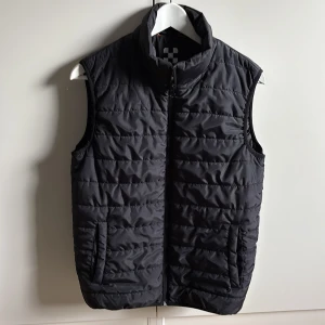 Svart dunväst - Snygg svart dunväst med dragkedja framtill och fickor på sidorna. Jättefint skick, ser oanvänd ut och har inga defekter. 