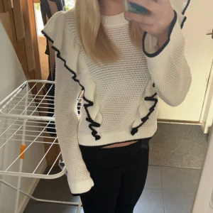 Vit stickad tröja med volangdetaljer - Säljer en snygg vit stickad tröja med svarta volangdetaljer längs ärmarna. Tröjan har långa ärmar och en bekväm passform. Säljer pga att den inte används längre💗