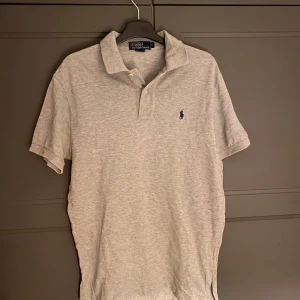Grå pikétröja från Ralph Lauren - Säljer en klassisk grå pikétröja från Ralph Lauren med den ikoniska loggan broderad på bröstet. Tröjan har korta ärmar och en stilren krage med knappar. Perfekt för en avslappnad men stilfull look.