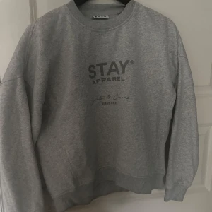 Grå sweatshirt från Stay - Säljer en grå sweatshirt från Stay med tryck på framsidan. Tröjan har en rund halsringning och långa ärmar. Perfekt för en avslappnad stil.