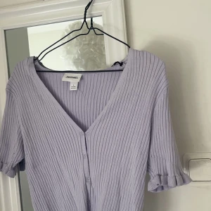 Ljuslila ribbad topp från Monki - Säljer en ljuslila ribbad topp från Monki med v-ringning och knappar framtill. Toppen har korta ärmar med volangdetaljer vid ärmsluten. Perfekt för en stilren look!