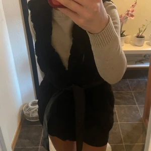 Svart pälsväst med knyt - Säljer en svart pälsväst med knytband i midjan. Västen är mjuk och fluffig, perfekt för att hålla sig varm och stilren under vårens dagar. Passar bra över en tröja eller klänning för en trendig look.