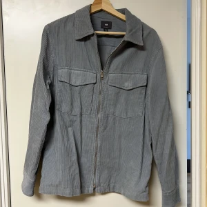 Grå manchesterjacka från H&M - Säljer en stilren grå manchesterjacka från H&M. Jackan har en dragkedja framtill och två bröstfickor med lock. Perfekt för en avslappnad look.