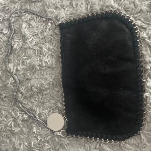 Svart axelväska med kedjedetaljer - Snygg svart axelväska med kedjedetaljer och dragkedja. Väskan har en rund metallbricka som dekoration och är perfekt för att bära dina nödvändigheter. Den har en stilren design med en kedja som axelrem.