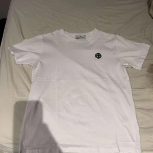 Vit t-shirt från Stone Island - Säljer en stilren vit t-shirt från Stone Island med deras ikoniska logga på bröstet. Perfekt för en casual look. T-shirten har en klassisk passform och är kortärmad.