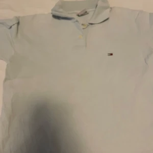 Ljusblå pikétröja från Tommy Hilfiger - Säljer en stilren ljusblå pikétröja från Tommy Hilfiger Denim. Tröjan har en klassisk krage och knappar framtill. Perfekt för en avslappnad men ändå snygg look.