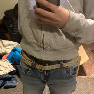 Beige flätat bälte - Säljer ett snyggt beige flätat bälte med bruna skinn-detaljer. Perfekt för att ge en stilren touch till din outfit. Bältet har en klassisk design och passar till många olika stilar.