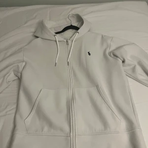 Vit kofta från Ralph Lauren - Säljer en stilren vit kofta från Ralph Lauren med dragkedja och det klassiska logotypen broderad på bröstet. Tröjan har långa ärmar och en praktisk huva med snörning. Perfekt för en avslappnad look.