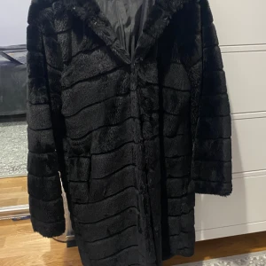 Svart pälsjacka med huva - Säljer en supermjuk och varm svart pälsjacka med huva. Perfekt för kalla vinterdagar! Jackan har långa ärmar och en stilren design utan synliga knappar eller dragkedja. Den är i mycket bra skick och redo för nya äventyr. Passar perfekt till både vardag och fest!