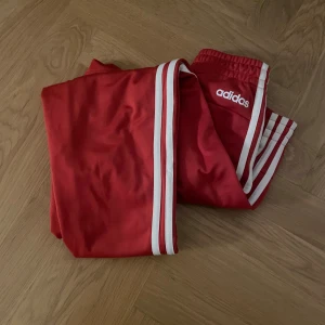 Röda träningsbyxor från Adidas - Säljer ett par snygga röda träningsbyxor från Adidas i storlek XS. Byxorna har de klassiska vita ränderna längs sidorna och en bekväm resårmidja. Perfekta för träning eller en avslappnad dag. 🏃‍♀️