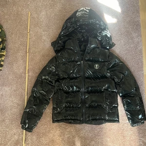 Svart pufferjacka från Trapstar - Säljer en snygg svart pufferjacka från Trapstar. Den är i mycket bra skick och perfekt för vintern. Jackan har en glansig finish med dragkedja och en stor huva. Den är långärmad och har en cool logga på bröstet. Perfekt för att hålla sig varm och stilren i kylan!
