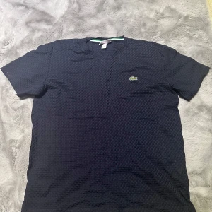 Mörkblå t-shirt från Lacoste - Säljer en snygg mörkblå t-shirt från Lacoste i mycket bra skick. Den har ett diskret rutmönster och det klassiska krokodilemblem på bröstet. Perfekt för både vardag och fest. Passar bra till jeans eller shorts!