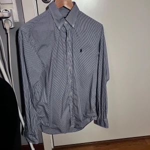 Randig skjorta från Ralph Lauren - Säljer en klassisk randig skjorta från Ralph Lauren i blått och vitt. Skjortan har långa ärmar och knappar framtill. Perfekt för både vardag och festliga tillfällen. Den är i mycket bra skick och gjord av bomull för en bekväm passform.