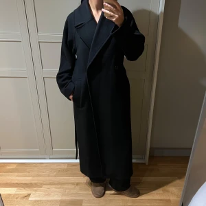 Svart lång kappa från Weekday - Säljer min oversized kappa från Weekday. Modellen heter Kia och är dubbelknäppt med långa ärmar, ett bälte i midjan för en snygg siluett samt en innerficka. Den är tillverkad i en mjuk, tovad ullblandning (59% ull, 29% polyester) med foder (100% polyester) vilket gör den varm nog för både höst och vinter. Jag har endast använt den en vinter. Den är i mycket bra skick utan några skador. Jag är 170 cm och storlek S sitter normalt. Nypris: 1500 kr.