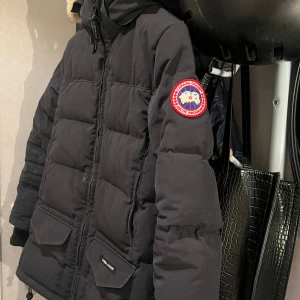 Svart dunjacka från Canada Goose - Säljer en svart Canada Goose dunjacka i mycket bra skick. Jackan har en fluffig pälsdetalj på huvan och är perfekt för kalla vinterdagar. Den har långa ärmar och praktiska fickor framtill. Passar perfekt för vinterns alla äventyr! ❄️