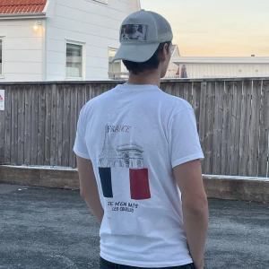 Vit t-shirt med franskt motiv - Säljer en vit t-shirt med tryck av Eiffeltornet och Triumfbågen på baksidan. Texten 'FRANCE' och 'JE M'EN BATS LES COUILLES' finns också. Framsidan har en liten logga 'N.' på bröstet. Perfekt för en avslappnad stil!