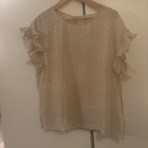 Beige blus med volangärmar - Säljer en söt beige blus med glittriga detaljer och volangärmar. Perfekt för både vardag och fest! Blusen har en lös passform och är i mycket bra skick. Den är kortärmad och har ett diskret mönster som ger en extra touch.