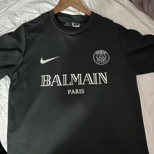 Svart fotbollströja från Nike x Balmain - Säljer en snygg svart fotbollströja från Nike i samarbete med Balmain. Tröjan har korta ärmar och ett stilrent tryck med Balmain Paris-loggan på bröstet. Perfekt för både träning och vardag. Passa på att köpa denna unika tröja!