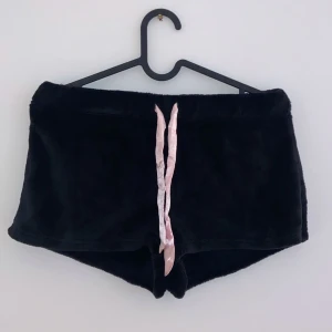 Svarta fluffiga shorts med kattmotiv - Supermysiga svarta shorts i fluffigt material med ett gulligt kattmotiv på baksidan. De har en rosa snörning framtill och texten 'MEOOW' broderad ovanför en liten hjärtdetalj. Perfekta för en chill dag hemma eller som en rolig present till kattälskaren! 🐱