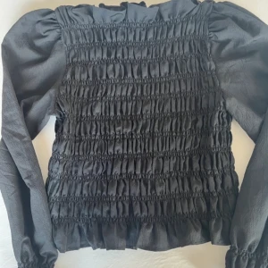 Svart smockad blus - Snygg svart blus med smockdetaljer som ger en cool textur. Den har långa ärmar och en elastisk passform som gör den både bekväm och stilren. Perfekt för både vardag och fest. Från hm i storlek S