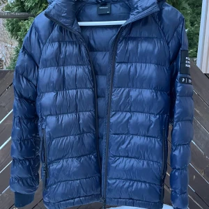 Blå dunjacka från Peak Performance - Säljer en snygg blå dunjacka från Peak Performance. Jackan har en dragkedja framtill och en praktisk huva. Perfekt för kyliga dagar med sin quiltade design som håller värmen. Den är i mycket bra skick och passar perfekt för höst och vinter.