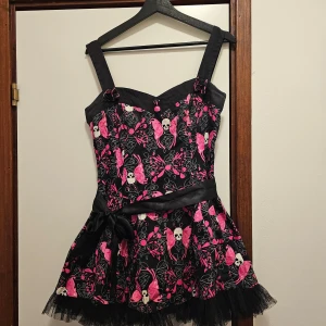 Svart och rosa klänning från Hell Bunny - Säljer en supercool svart klänning från Hell Bunny med rosa och vita dödskallemönster. Den har breda axelband och en söt rosett i midjan. Kjolen är kort och har en volangkant i tyll. Perfekt för en fest eller Halloween! 🖤💀 Axelbanden har två knapphål så man kan ändra längden. 