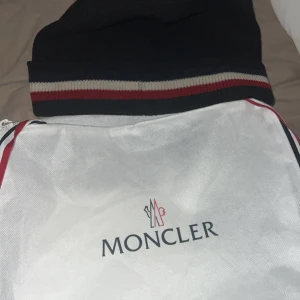Svart mössa från Moncler - Säljer en snygg svart mössa från Moncler med röd och vit randdetalj. Perfekt för att hålla sig varm och stilren under kyliga dagar.  och kommer med originalförpackning.