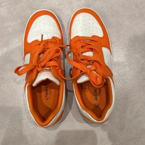 Vita och orange sneakers från Monki - Säljer ett par snygga vita och orange sneakers från Monki. De har en cool design med snörning och är i bra skick. Perfekta för en casual look eller för att liva upp en outfit med lite färg!