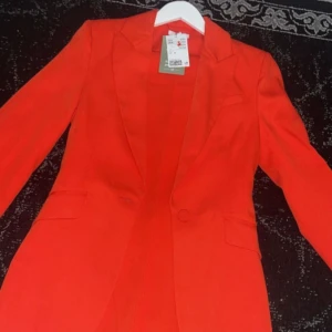 Röd kavaj - Säljer en snygg röd kavaj i nytt skick. Perfekt för att liva upp garderoben! Den har en klassisk design med långa ärmar och en enkel knapp framtill. Passar både till vardags och fest. Storlek är inte angiven, men den ser ut att ha en normal passform. Byxor 36 och kavaj 34.