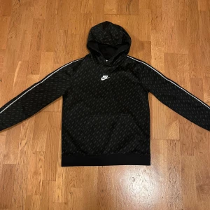 Svart hoodie från Nike - Säljer en svart hoodie från Nike i storlek L. Den har ett diskret mönster och en vit Nike-logga på bröstet. Hoodien har långa ärmar med vita ränder längs sidorna och en praktisk magficka. Perfekt för både träning och vardag!