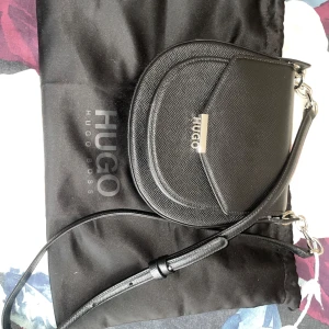 Svart axelväska från Hugo Boss - Snygg och stilren svart axelväska från Hugo Boss i mycket bra skick. Väskan har en elegant design med silverdetaljer och justerbar axelrem. Perfekt för både vardag och fest!