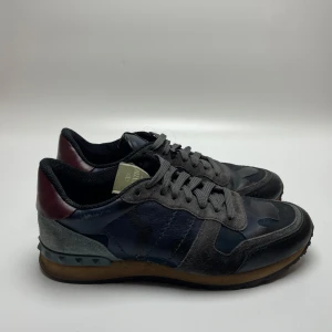 Valentino Rockrunner i camouflage - Säljer ett par snygga Valentino Rockrunner sneakers i en cool camouflage-design. Skorna har en mix av grått och svart med detaljer i mörkrött och en gummisula. De är i bra skick och perfekta för både vardag och fest. Storlek är inte specificerad, men de ser ut att passa en normal passform. 39 passar 40