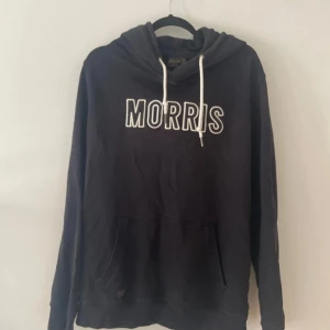 Svart hoodie från Morris - Säljer en snygg svart hoodie från Morris i storlek XL. Den har en klassisk design med vit text på bröstet och justerbara snören i huvan. Perfekt för en avslappnad stil och passar till alla tillfällen. Den är i bra skick och redo för en ny ägare!