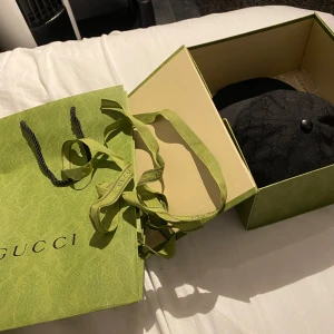 Svart keps från Gucci - Säljer en snygg svart keps från Gucci med deras ikoniska mönster. Kepsen har en röd och grön rand på sidan och kommer i originalförpackning. Perfekt för att ge din outfit en lyxig touch. Passar både till vardags och fest!