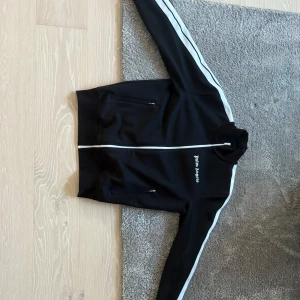 Svart träningsjacka från Palm Angels - Säljer en snygg svart träningsjacka från Palm Angels i storlek M. Jackan har vita ränder längs ärmarna och en dragkedja framtill. Perfekt för både träning och vardagsbruk. Den är i mycket bra skick och gjord av polyester för en bekväm passform.