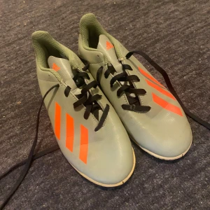 Gröna fotbollsskor från Adidas - Säljer ett par gröna Adidas fotbollsskor med orangea detaljer. Skorna har snörning och är i bra skick. Perfekta för fotbollsträning eller match. De har en snygg och modern design med de klassiska tre ränderna på sidan.