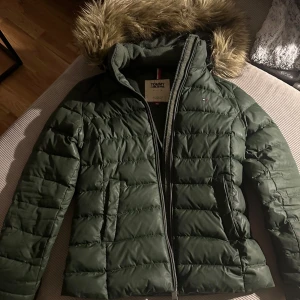 Grön dunjacka från Tommy Hilfiger - Säljer en snygg grön dunjacka från Tommy Hilfiger med pälskantad huva. Jackan har en dragkedja framtill och är perfekt för kalla vinterdagar. Den är i mycket bra skick och har långa ärmar. Passar perfekt för både vardag och vinteraktiviteter.