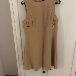 Beige ärmlös klänning från Warehouse - Snygg beige ärmlös klänning från Warehouse i storlek 40. Klänningen har dekorativa knappar framtill och en dragkedja bak. Perfekt för både vardag och festliga tillfällen. Stilren och enkel design som passar till många olika tillfällen. Obs en knapp fattas, annars i mycket gott då jag använt den totalt två gånger. 