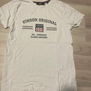 Säljer en vit t-shirt från Vinson Original i storlek M. Den har tryck på framsidan med texten 'Legendary Traditions' och 'All-American Classic Heritage'. Perfekt för en avslappnad stil. T-shirten är i bra skick och gjord av mjuk bomull.