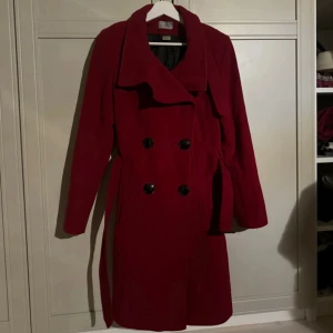 Burgundy Ullkappa  - Säljer en snygg röd kappa i mycket bra skick. Den har en klassisk dubbelknäppning med stora svarta knappar och ett bälte i midjan för en elegant look. Perfekt för höst och vinter, med långa ärmar och en stilren design. Passar både till vardags och festliga tillfällen. 80% ull 20% polyamid.  Bara lite slitet på spännena som man ser på bilden, men det syns knappt. Jättefin kappa 🌹