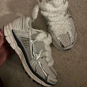 Nike vomero 5 Silver - Säljer ett par coola silvriga sneakers med unika fluffiga snören. De har en futuristisk design med meshdetaljer och en bekväm sula. Perfekta för att sticka ut i mängden och ge din outfit en extra touch. Passar både till vardags och fest!