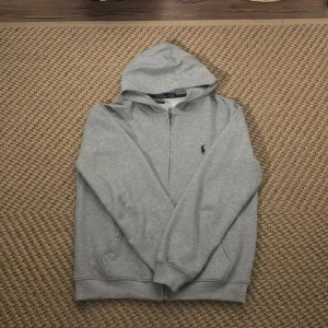 Grå hoodie från Polo Ralph Lauren - Säljer en snygg grå hoodie från Polo Ralph Lauren i storlek M. Den har en dragkedja framtill och en klassisk logga på bröstet. Perfekt för både höst och vår, och den är superbekväm med sin mjuka bomullskvalitet. Passar till alla tillfällen, från chill till casual outings.