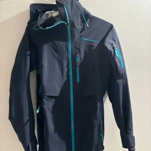 Mörkblå vindjacka från Peak Performance - Säljer en marinblå vindjacka/skidjacka från Peak Performance i mycket bra skick. Jackan har en snygg turkos dragkedja och flera praktiska fickor. Perfekt för höst och vår, med långärmad design och justerbar huva. Passar både för vardag och utomhusaktiviteter.