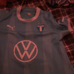 Ljusblå fotbollströja från Puma - Säljer en ljusblå fotbollströja från Puma med korta ärmar. Tröjan har ett randigt mönster och klubbmärke på bröstet. Den har även nummer 22 och namnet 'Ali' på ryggen. Perfekt för fotbollsfans! ⚽