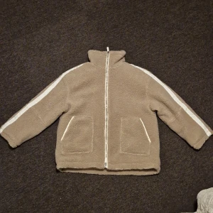 Beige teddyjacka med dragkedja - Mysig beige teddyjacka med dragkedja och vita detaljer längs ärmarna. Perfekt för kyliga dagar med sina stora fickor och höga krage. Jackan har en avslappnad passform och är superbekväm att bära. Perfekt för höst och vinter!