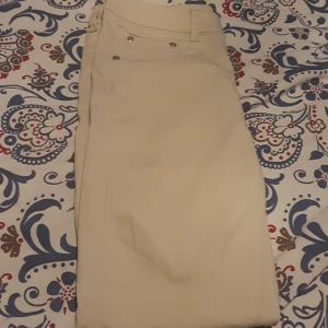 Beige byxor med fickor - Säljer ett par stilrena beige byxor i bra skick. De har en normal passform och är perfekta för både vardag och fest. Byxorna har praktiska fickor och knappar framtill. Materialet känns mjukt och bekvämt, vilket gör dem sköna att bära hela dagen. Passar perfekt till en avslappnad eller uppklädd stil! Finns en litet märke, se bilden. Byxorna är använda vid ett tillfälle.