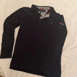 Marinblå  långärmad pikétröja från Tommy Hilfiger - Säljer en stilren marinblå långärmad pikétröja från Tommy Hilfiger. Den har klassiska knappar och en snygg randig detalj på insidan av kragen. Tröjan är i använt skick och ev lite urtvättad. Ca stl 150-160