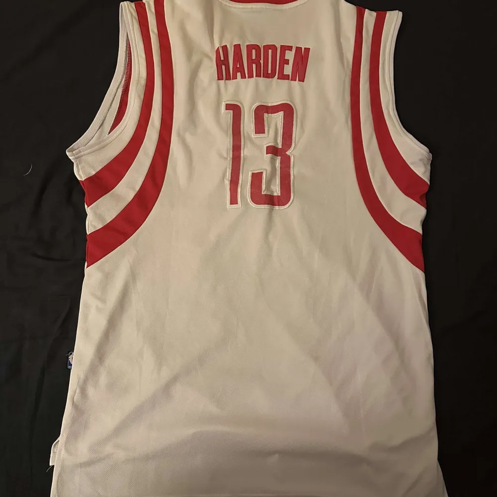 Säljer en vit och röd basketlinne från Adidas med texten 'Rockets' och nummer 13 på framsidan. Baksidan har namnet 'Harden' och samma nummer. Linnet är i bra skick och perfekt för basketfans. Det är ärmlöst och har ett sportigt utseende.. T-shirts.